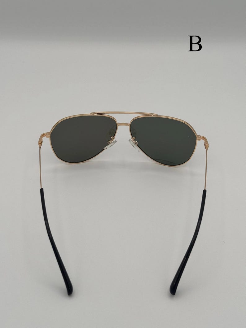 Puede incluir: Gafas de sol de aviador con montura dorada y lentes oscuras. Las gafas tienen un doble puente y patillas negras. La imagen est&aacute; sobre un fondo blanco.