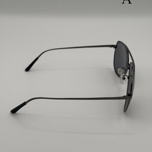 Puede incluir: Gafas de sol aviador de color gris oscuro con una fina montura met&aacute;lica. Las gafas de sol tienen una forma cl&aacute;sica de l&aacute;grima y lentes oscuras. Las patillas son delgadas y se curvan suavemente hacia los extremos. La letra "A" es visible.