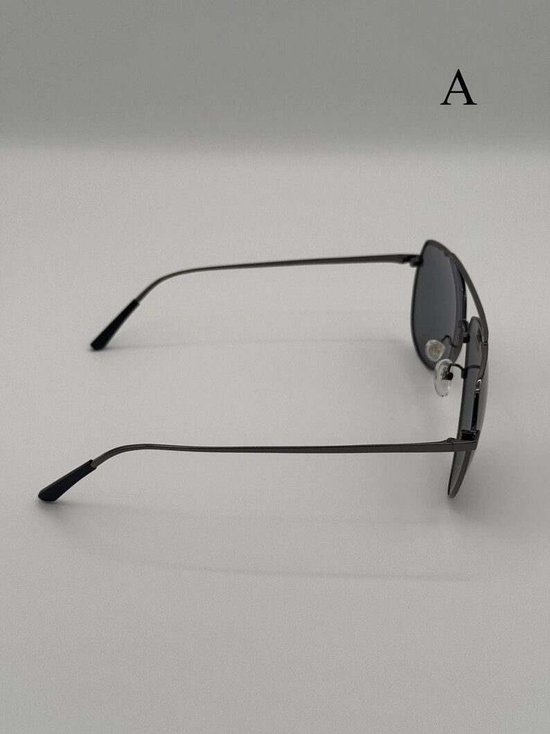 Puede incluir: Gafas de sol aviador de color gris oscuro con una fina montura met&aacute;lica. Las gafas de sol tienen una forma cl&aacute;sica de l&aacute;grima y lentes oscuras. Las patillas son delgadas y se curvan suavemente hacia los extremos. La letra "A" es visible.