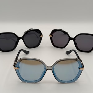 May include: Three pairs of sunglasses with a geometric design. Two pairs have black frames and lenses, with gold-colored accents. The third pair has a clear frame with blue lenses and gold-colored accents. All have black arms.