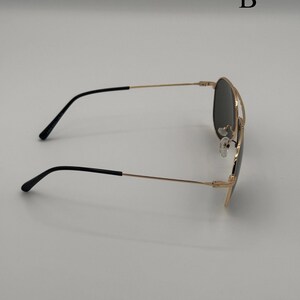 Puede incluir: Gafas de sol de aviador con montura dorada y lentes negras. Las gafas tienen un doble puente y patillas negras. La letra "B" es visible en el fondo.
