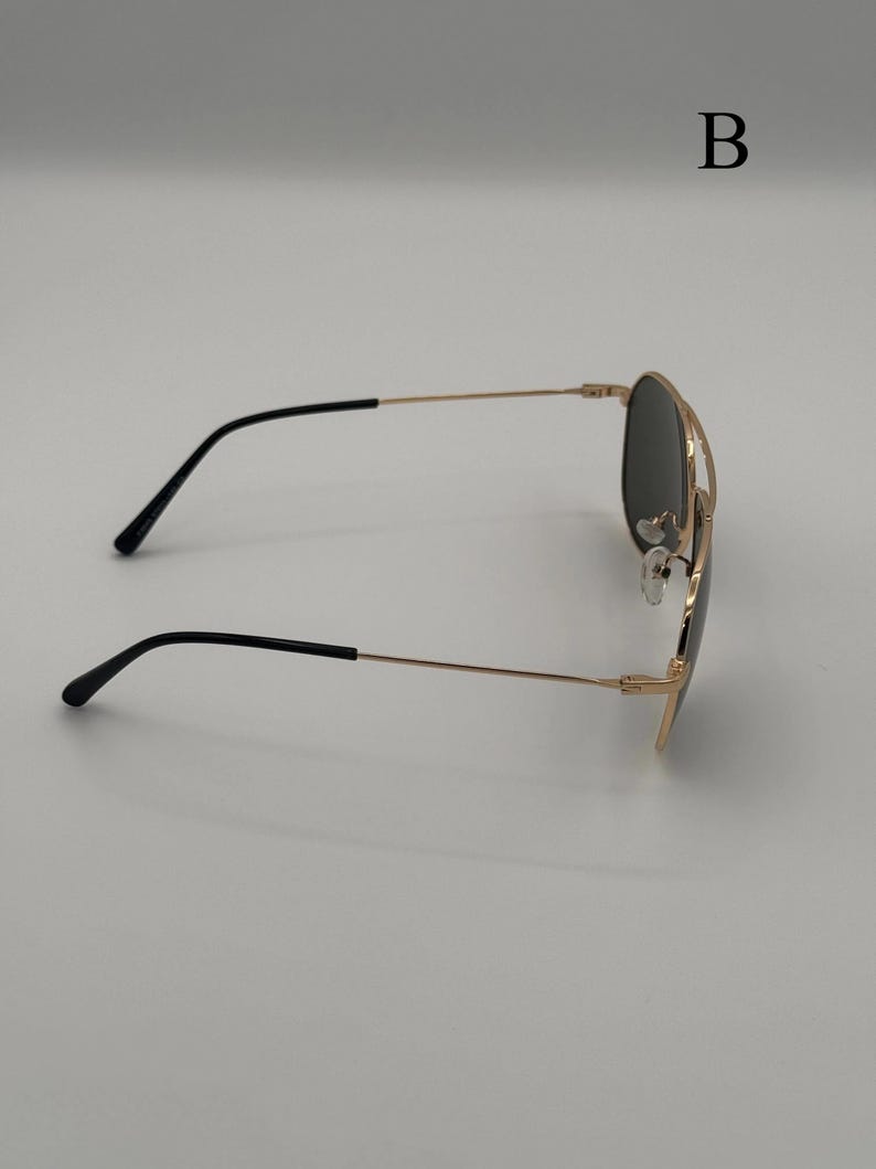 Puede incluir: Gafas de sol de aviador con montura dorada y lentes negras. Las gafas tienen un doble puente y patillas negras. La letra "B" es visible en el fondo.