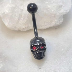 May include: A black belly button ring featuring a skull design. The skull has red gem eyes and is attached to a black curved barbell. The top ball is also black. The jewelry is shown against a white background.