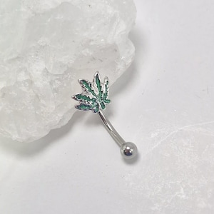 May include: A curved eyebrow ring with a silver-colored bar and a small, round ball at one end. The decorative element is a green and silver cannabis leaf design. The jewelry is set against a white background.