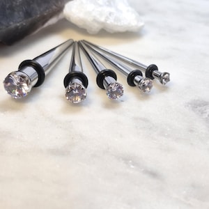 May include: A collection of silver-toned ear stretching tapers with clear, faceted gem ends. Each taper has a black rubber O-ring. The tapers are arranged on a white marble surface, with a dark gray stone and white rock in the background.