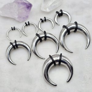 May include: A set of seven silver crescent moon shaped body jewelry pieces with black rubber o-rings. The jewelry is arranged in a semi-circle on a white surface.
