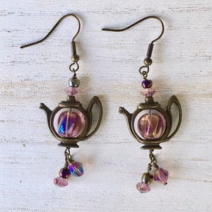 May include: A pair of dangle earrings featuring a teapot charm with a pink iridescent glass bead inside. The teapot is made of bronze metal and has a small bead at the top. The earrings are finished with a dangling pink iridescent glass bead.