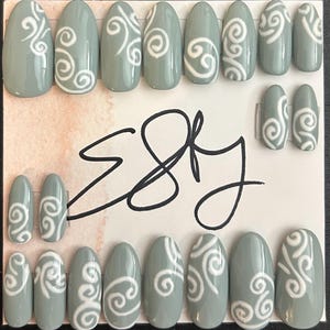 May include: A set of sage green press-on nails with a white swirl design. The nails are oval-shaped and arranged on a card with the word "Etsy" in a handwritten font.