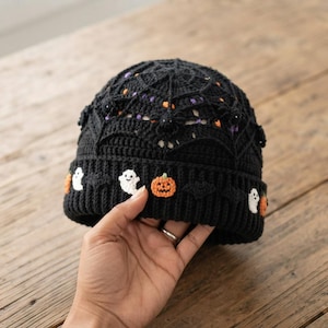 May include: A black crocheted beanie featuring a detailed spiderweb pattern on the crown. The ribbed cuff is decorated with small, colorful crocheted appliques including white ghosts, orange pumpkins, and black bats arranged in a repeating pattern.