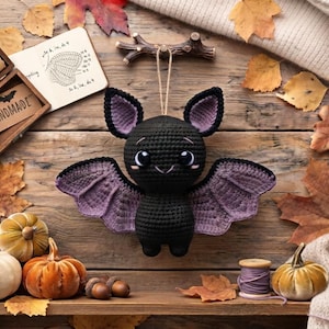 May include: A black and purple crocheted bat plush toy with large eyes, pointed ears, and outstretched wings, suspended by a string from a small wooden branch, featuring a detailed stitch pattern and a soft, rounded body design.