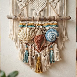 May include: A cream-colored macrame wall hanging suspended from a wooden branch, featuring three crocheted seashells in gold, brown, and blue. The piece is adorned with small white cowrie shells, wooden beads, and hanging tassels in blue and gold.