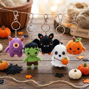 May include: Five crochet keychains hanging from a string, featuring a purple one-eyed monster, a green Frankenstein-style figure, a black bat, a white ghost holding a small orange pumpkin, and an orange pumpkin with a green stem.