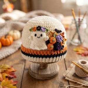 May include: A cream-colored crochet beanie featuring a ghost appliqué with a floral crown, accented by a cluster of crocheted pumpkins and leaves. The brim has horizontal stripes in orange and black, creating a seasonal autumn-themed accessory.