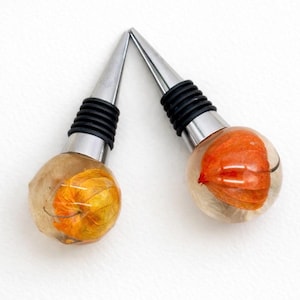 Könnte beinhalten: Zwei dekorative Flaschenverschlüsse. Jeder Verschluss hat eine klare, abgerundete Harzkugel mit einer orangefarbenen und gelben getrockneten Blume im Inneren. Die Verschlüsse haben eine silberfarbene, konische Spitze und einen schwarzen Gummisockel.