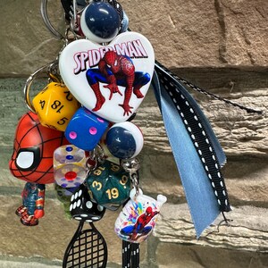 Pode incluir: Uma cole&ccedil;&atilde;o de chaveiros e berloques com o tema Homem-Aranha. Inclui um berloque em forma de cora&ccedil;&atilde;o com o logotipo do Homem-Aranha, dados, um berloque de raquete de t&ecirc;nis e uma pequena figura do Homem-Aranha. Os chaveiros est&atilde;o presos a uma fita azul.