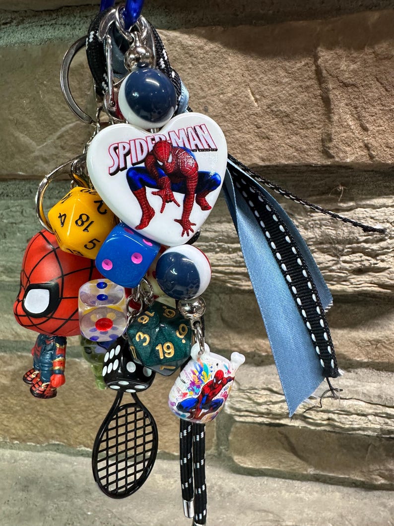 Pode incluir: Uma cole&ccedil;&atilde;o de chaveiros e berloques com o tema Homem-Aranha. Inclui um berloque em forma de cora&ccedil;&atilde;o com o logotipo do Homem-Aranha, dados, um berloque de raquete de t&ecirc;nis e uma pequena figura do Homem-Aranha. Os chaveiros est&atilde;o presos a uma fita azul.