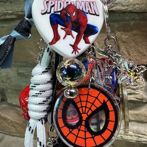 May include: A selection of Spider-Man themed keyrings and charms. The keyrings feature a heart-shaped charm with the Spider-Man logo and a graphic of Spider-Man, a round charm with the Spider-Man mask, and other charms.