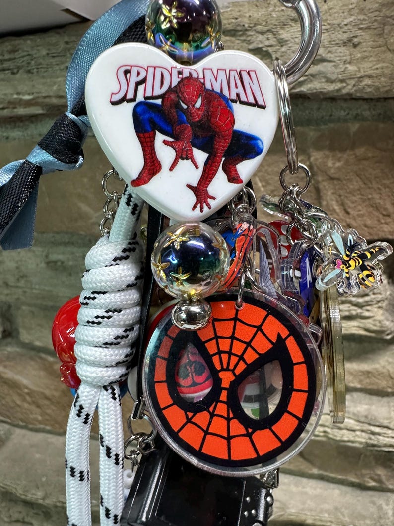 May include: A selection of Spider-Man themed keyrings and charms. The keyrings feature a heart-shaped charm with the Spider-Man logo and a graphic of Spider-Man, a round charm with the Spider-Man mask, and other charms.