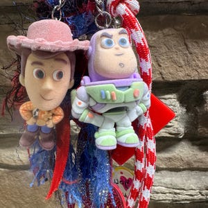 May include: A keychain featuring two character figures: Woody in a pink hat and Buzz Lightyear in a space suit. The keychains are attached to a chain and colourful decorative elements, including red and blue ribbons.