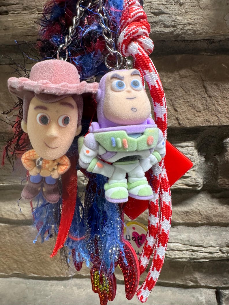 May include: A keychain featuring two character figures: Woody in a pink hat and Buzz Lightyear in a space suit. The keychains are attached to a chain and colourful decorative elements, including red and blue ribbons.