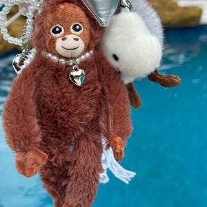 Peut inclure: Un porte-clés orang-outan en peluche marron avec un collier de perles et un pendentif en forme de cœur. Un pompon blanc avec une queue marron et un nœud argenté sont attachés. L'orang-outan a les mains et les pieds oranges. Une chaîne perlée avec des breloques en forme de cœur est également attachée.