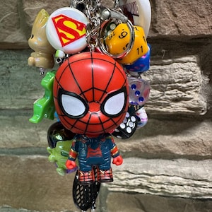 Pode incluir: Uma cole&ccedil;&atilde;o de chaveiros com v&aacute;rios personagens da cultura pop. O foco principal &eacute; uma figura vermelha e preta do Homem-Aranha. Outros amuletos incluem um logotipo do Superman, um dado amarelo e outras figuras. Os chaveiros s&atilde;o presos a uma corrente prateada.