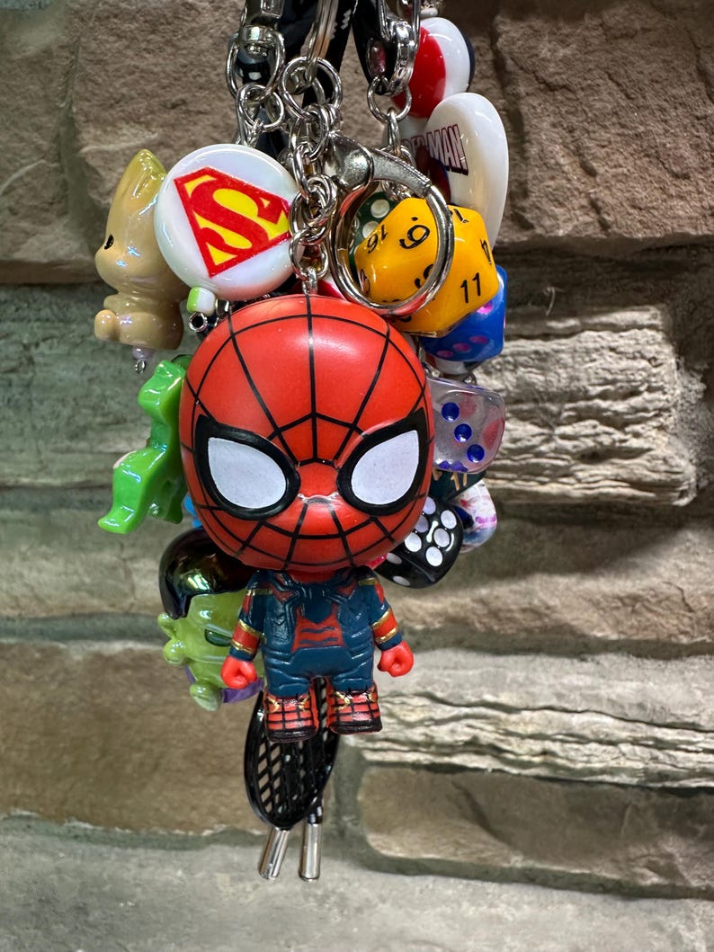 Pode incluir: Uma cole&ccedil;&atilde;o de chaveiros com v&aacute;rios personagens da cultura pop. O foco principal &eacute; uma figura vermelha e preta do Homem-Aranha. Outros amuletos incluem um logotipo do Superman, um dado amarelo e outras figuras. Os chaveiros s&atilde;o presos a uma corrente prateada.