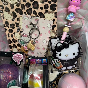 Könnte beinhalten: Eine Sammlung von Hello Kitty Artikeln, bestehend aus einem Notizbuch mit Leopardenmuster, einem Metallschlüsselanhänger mit Anhänger, einem rosa Stift mit Figurenaufsatz, einem lila glitzernden Squishy und einem Nee Doh Peace O' Cake Spielzeug.