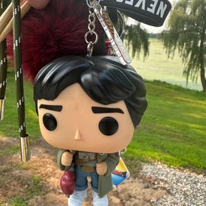 May include: A keychain featuring a stylized vinyl figure with black hair, a green coat, blue pants, and white shoes. The figure holds a red boxing glove. Attached are a dark red pom-pom, a black strap with the text NEVER SAY DIE, and a clear charm.