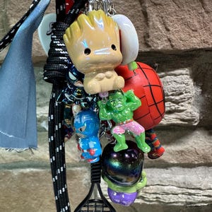 Pode incluir: Um porta-chaves com figuras de personagens, incluindo Groot, Homem-Aranha, Hulk, Capit&atilde;o Am&eacute;rica e um pingente de raquete de t&ecirc;nis. Os porta-chaves est&atilde;o presos a um porta-chaves prateado com um cord&atilde;o preto e azul.