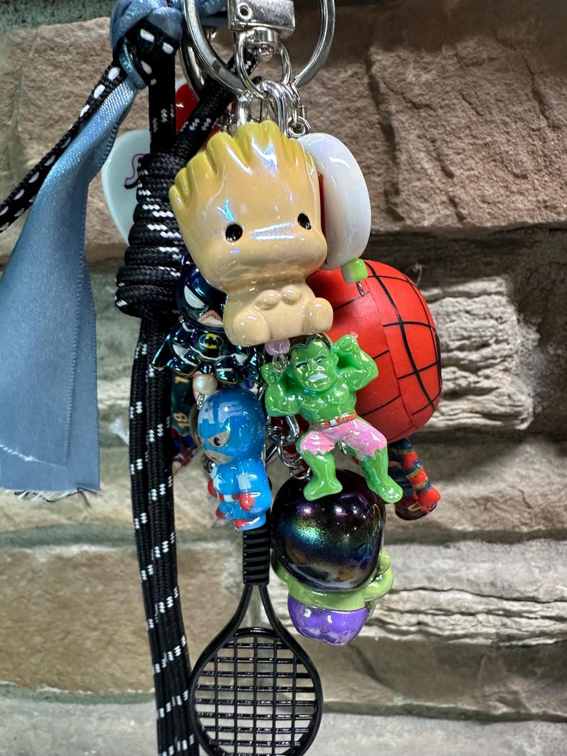 Pode incluir: Um porta-chaves com figuras de personagens, incluindo Groot, Homem-Aranha, Hulk, Capit&atilde;o Am&eacute;rica e um pingente de raquete de t&ecirc;nis. Os porta-chaves est&atilde;o presos a um porta-chaves prateado com um cord&atilde;o preto e azul.