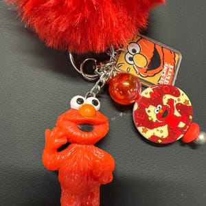 May include: A red Elmo keychain with a fluffy red pom-pom. The keychain includes an Elmo figurine, a Sesame Street tag, and a round charm with Elmo's face. The charms are attached to a silver chain.