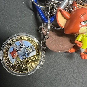 May include: A selection of Zootopia-themed keyrings and accessories. Features a clear, round coin with the Zootopia logo and characters, a fox figurine, and various metal keyrings and blue carabiner clips. The coin reads "Zootopia Welcomes New Zootizens."