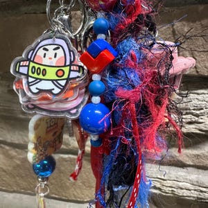 May include: A keychain featuring a cartoon character charm, blue and red beads, and a cluster of red, blue, and black fibres. The keychain includes a silver clasp and a charm with the text "Toy Story".