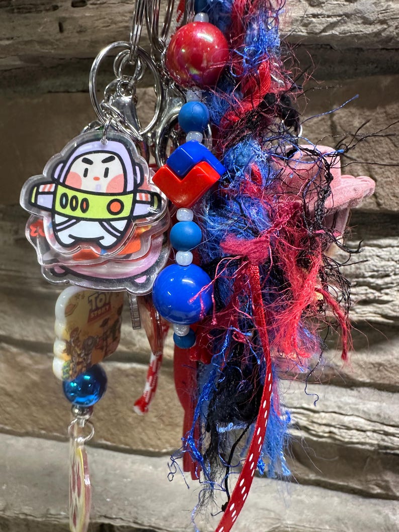 May include: A keychain featuring a cartoon character charm, blue and red beads, and a cluster of red, blue, and black fibres. The keychain includes a silver clasp and a charm with the text "Toy Story".