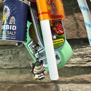 May include: A keychain featuring multiple charms including a blue Morbid Dying Salt container, a Heatwave Dave acrylic charm, a white and orange cigarette-shaped charm, and a small figure wearing a black helmet and white suit with a backpack.