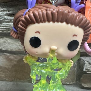 May include: A vinyl figure keychain featuring a character with brown hair in pigtails, black eyes, and a purple outfit. The figure has a translucent, neon green slime effect dripping from its nose and hands, attached to a purple plastic clip.