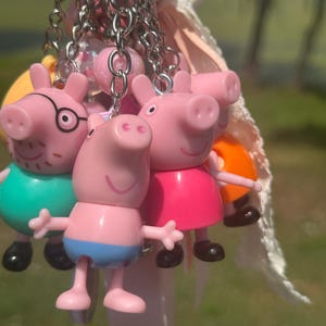 Pode incluir: Um porta-chaves com várias figuras de porquinhos cor-de-rosa. Uma figura veste uma camisola azul-petróleo, outra um vestido cor-de-rosa vivo e uma terceira calções azuis. As figuras estão presas a correntes de metal prateado com missangas e uma fita branca.