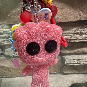 May include: A keychain featuring a pink, glittery, humanoid figure with large black eyes. The keychain includes a red cord, a red heart-shaped charm, a small yellow charm, and a heart-shaped pendant with a blue figure and text on a beach background.