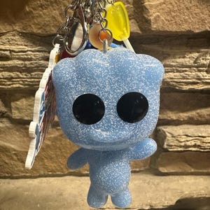 May include: A keychain featuring a light blue, glittery, humanoid figure with large black circular eyes. The figure is attached to a metal ring along with a yellow lollipop charm and a flat, multi-colored acrylic charm hanging from silver chains.