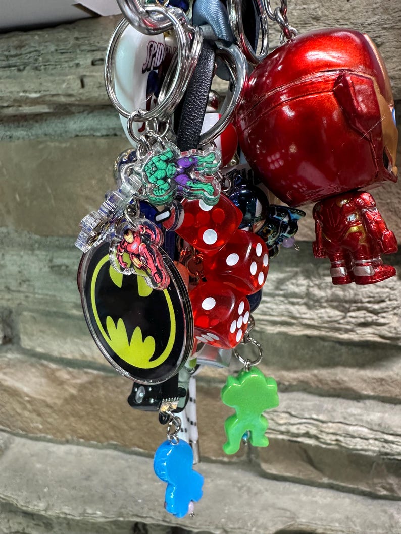 May include: A collection of keyrings with various designs. Includes a red Iron Man figure, Batman logo, Hulk, and red dice. Other keyrings have a blue, green, and white colour scheme. The keyrings are attached to silver keyrings.