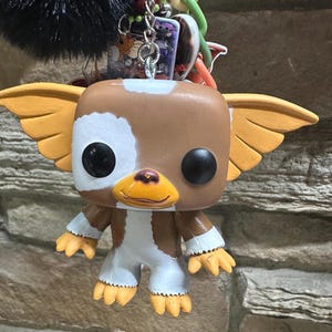 May include: A keychain featuring a brown and white Gizmo character figure with large ears and black eyes, attached to a silver metal ring with a black pom-pom, a green plastic chain, and several small decorative charms including a rectangular graphic piece.