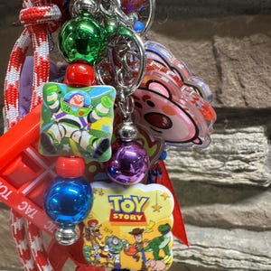 May include: A vibrant keychain with multiple charms and beads. It features a Buzz Lightyear square charm, a Toy Story logo charm, and a pink bear charm. The keychain also has red, green, blue, and purple beads, and a red and white striped lanyard.