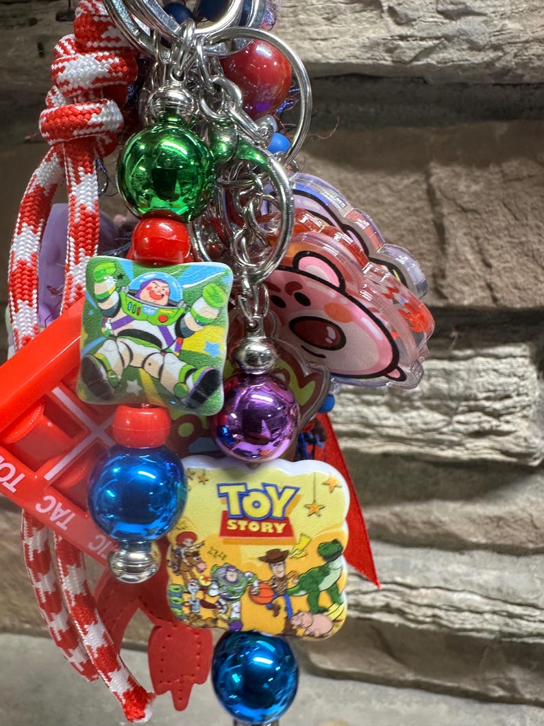 May include: A vibrant keychain with multiple charms and beads. It features a Buzz Lightyear square charm, a Toy Story logo charm, and a pink bear charm. The keychain also has red, green, blue, and purple beads, and a red and white striped lanyard.