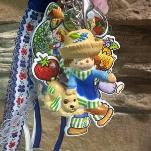 Può includere: Un portachiavi con una figura tridimensionale di un bambino con cappello di paglia e un cagnolino, accompagnato da ciondoli piatti in acrilico a forma di fragola e casa, fissato a un cordino intrecciato blu e un nastro bianco a fiori.