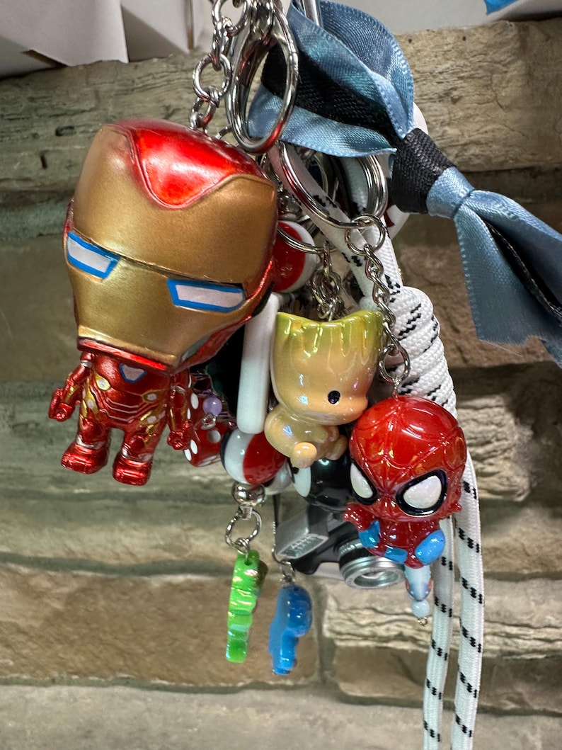 May include: A selection of superhero keyrings featuring Iron Man, Spider-Man, and Baby Groot characters, plus other colourful charms. The keyrings are attached to metal rings and a lanyard with a blue ribbon.