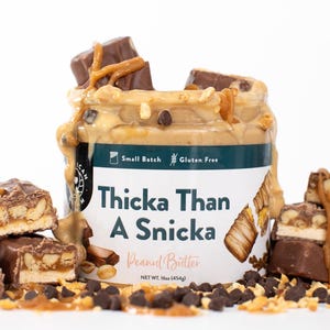 May include: A jar of peanut butter labelled "Thicka Than A Snicka", overflowing with peanut butter, chocolate chips, and candy bar pieces. The label also states "Small Batch" and "Gluten Free". Net weight is 454g.