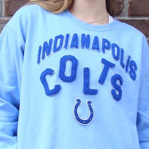 May include: Pale blue sweatshirt with the text "INDIANAPOLIS COLTS" in a dark blue, textured font. A dark blue horseshoe emblem is below the text. The sweatshirt has long sleeves and a crew neck.