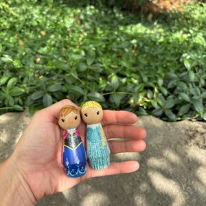May include: Two wooden peg dolls, one with brown hair and a blue and black dress, the other with blonde hair and a blue dress. The dolls are held in a hand against a background of green foliage.