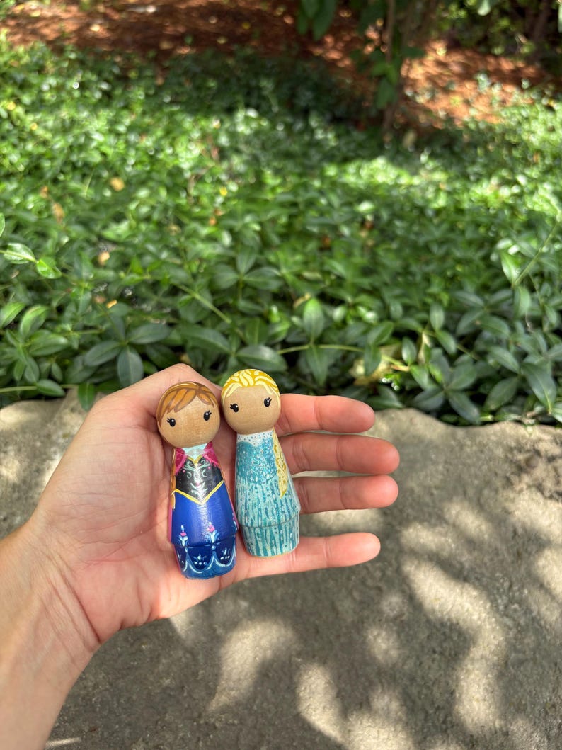 May include: Two wooden peg dolls, one with brown hair and a blue and black dress, the other with blonde hair and a blue dress. The dolls are held in a hand against a background of green foliage.